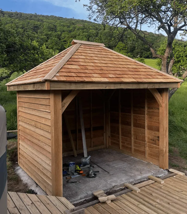 Open-front timber garden building with oak braces and a shingle roof